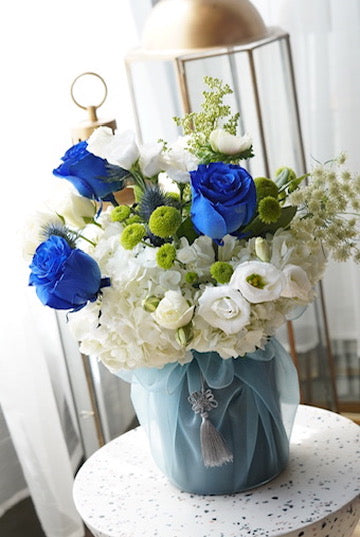 Mother's Day flower arrangement featuring vibrant blue roses, white ranunculus, and green pompon chrysanthemums in a blue fabric-wrapped vase with a traditional silver Korean tassel. Luxury floral delivery in Toronto and Oakville.
