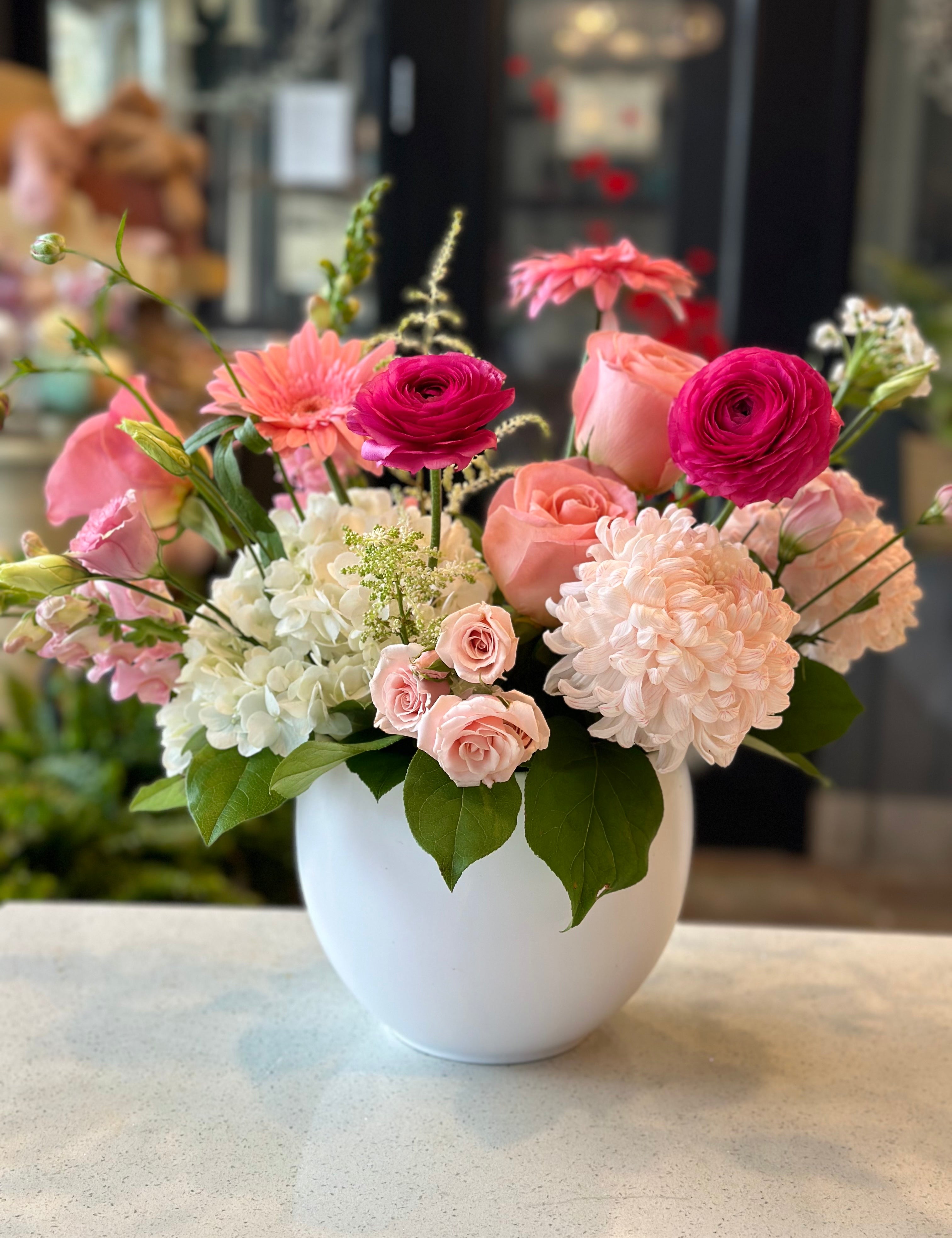 A floral arrangement featuring a variety of flowers including pink roses and hydrangeas, in a white vase, with a blurred background.