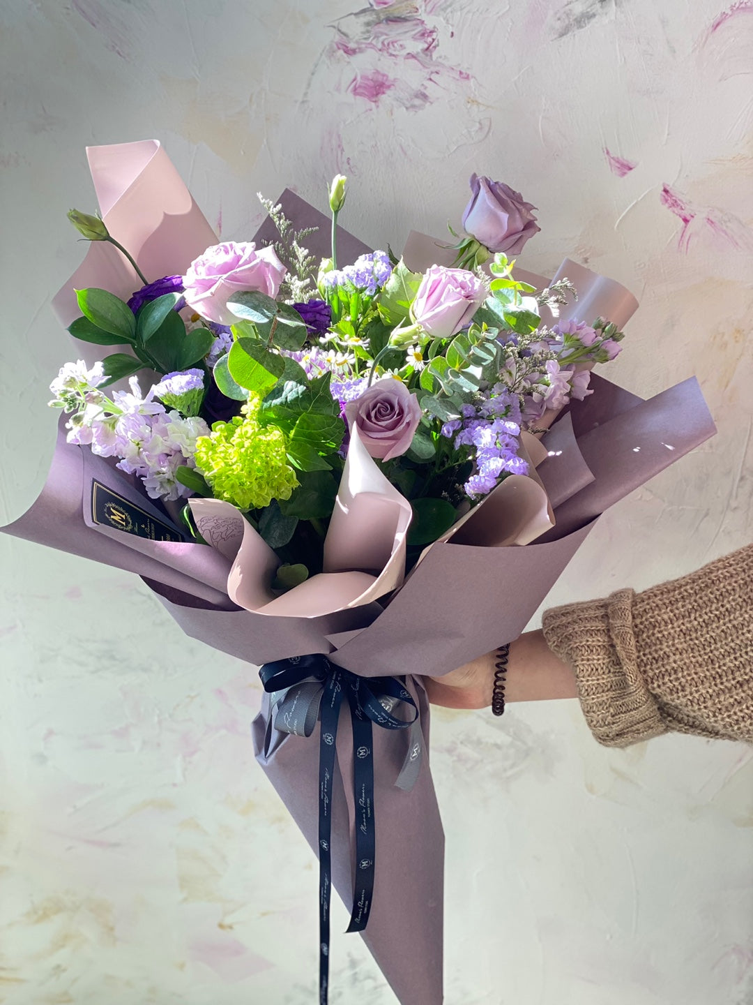 A person holding the Purple Rush Mother's Day bouquet, showing its generous size and vibrant purple roses, lisianthus, and green accents. Professional hand-tied floral gift by Moon's Flowers Toronto