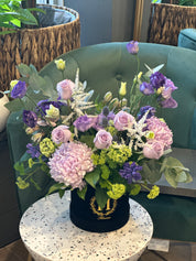 A floral arrangement with purple, green, and white flowers in a round black container, placed on a round white table.