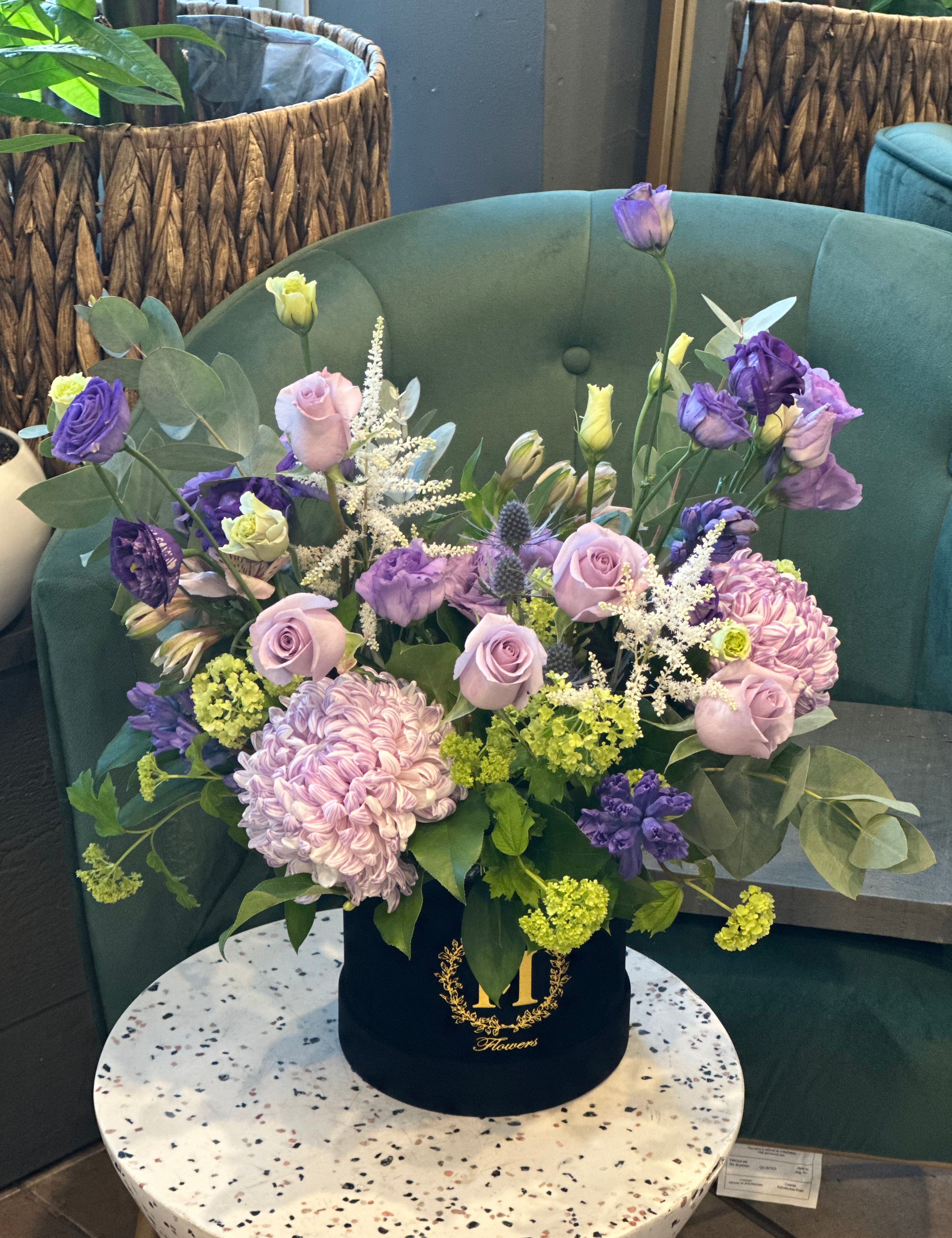 A floral arrangement with purple, green, and white flowers in a round black container, placed on a round white table.