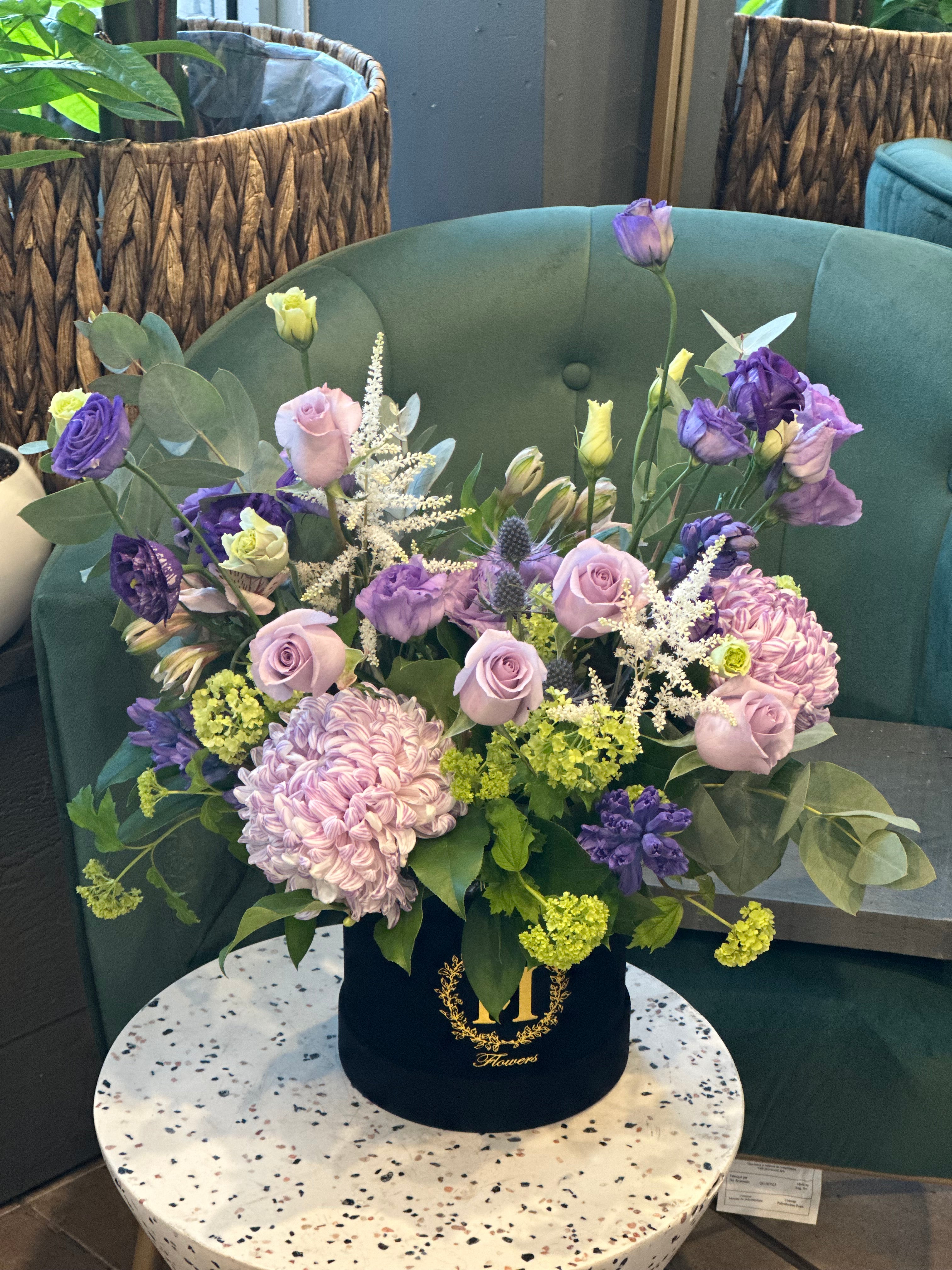 A floral arrangement with purple, green, and white flowers in a round black container, placed on a round white table.