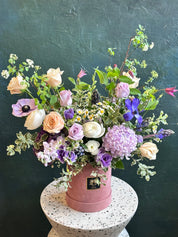 Pink Blossom Box in a pink box with fresh purple blooms like anemone, peach roses, and other greenery and filler like chamomile. 