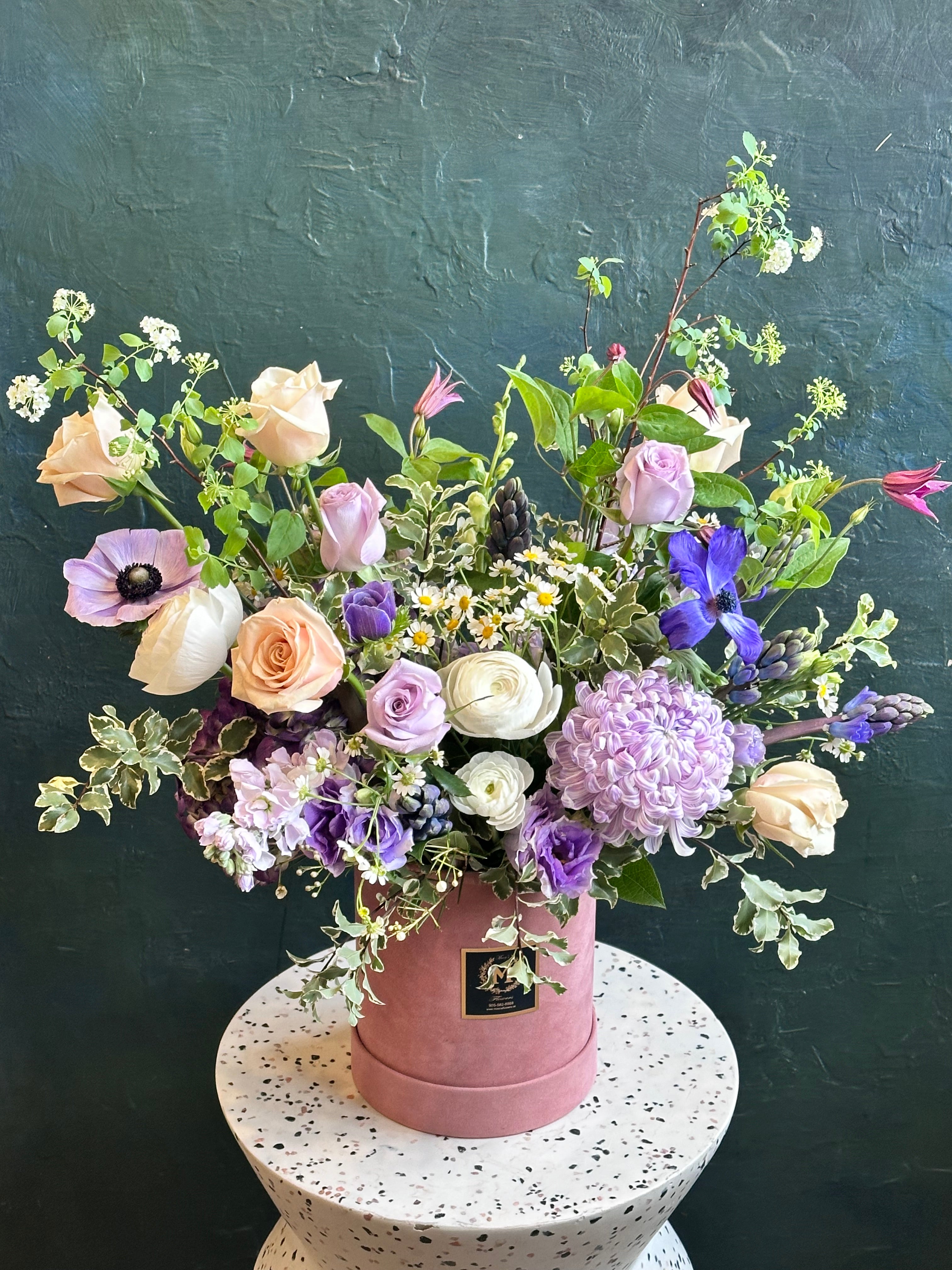 Pink Blossom Box in a pink box with fresh purple blooms like anemone, peach roses, and other greenery and filler like chamomile. 