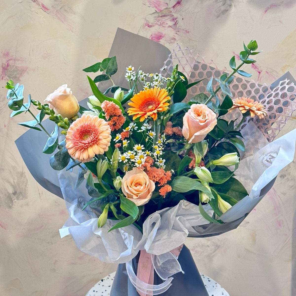 A bouquet of flowers including peach roses, orange gerberas, white lilies, and chamomile daisies, wrapped in gray paper with a peach ribbon.
