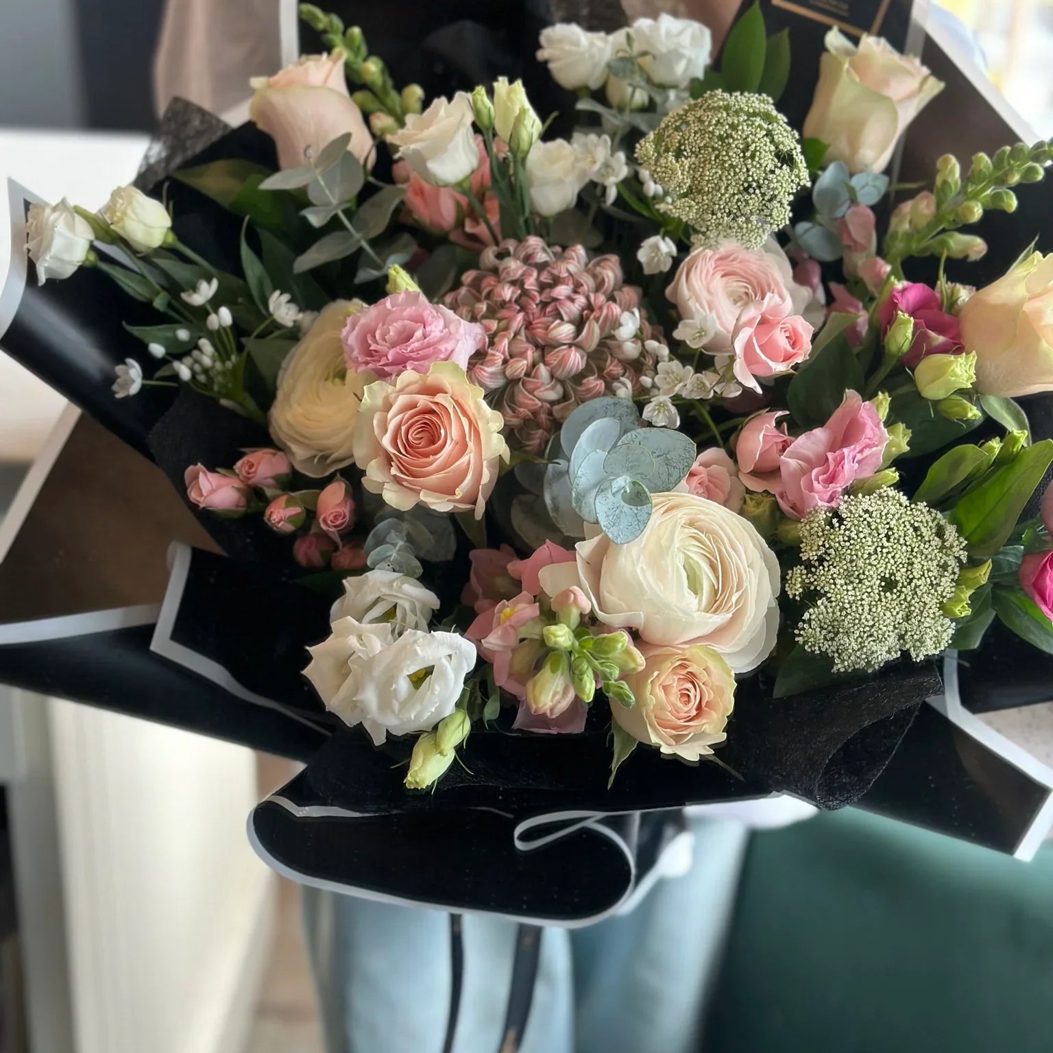 Modern and moody floral design featuring pastel-toned roses and textured greenery wrapped in bold black paper. Elegant 'Florist's choice' bouquet with high contrast styling by Moon's Flowers. Available for luxury flower delivery in Oakville and Toronto