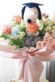 Snoopy graduation bouquet full view with pastel flowers, blue hydrangea, peach roses, and pink wrapping