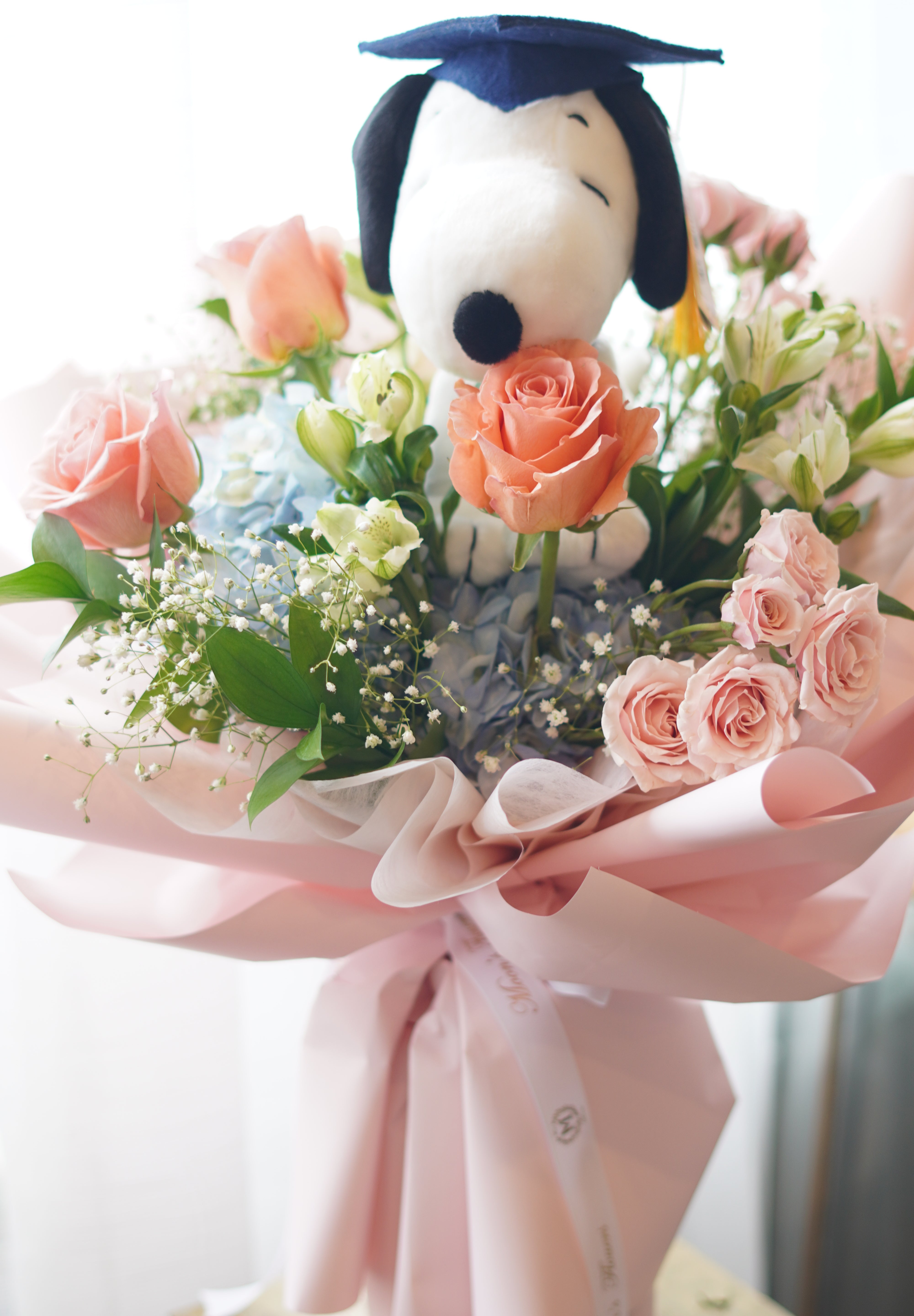 Snoopy graduation bouquet full view with pastel flowers, blue hydrangea, peach roses, and pink wrapping