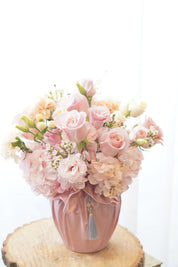 Soft pink and peach Mother's Day floral arrangement with roses and lisianthus in a pink fabric-wrapped vase on a rustic wood slice. Elegant flower delivery in Toronto and Oakville by Moon's Flowers.