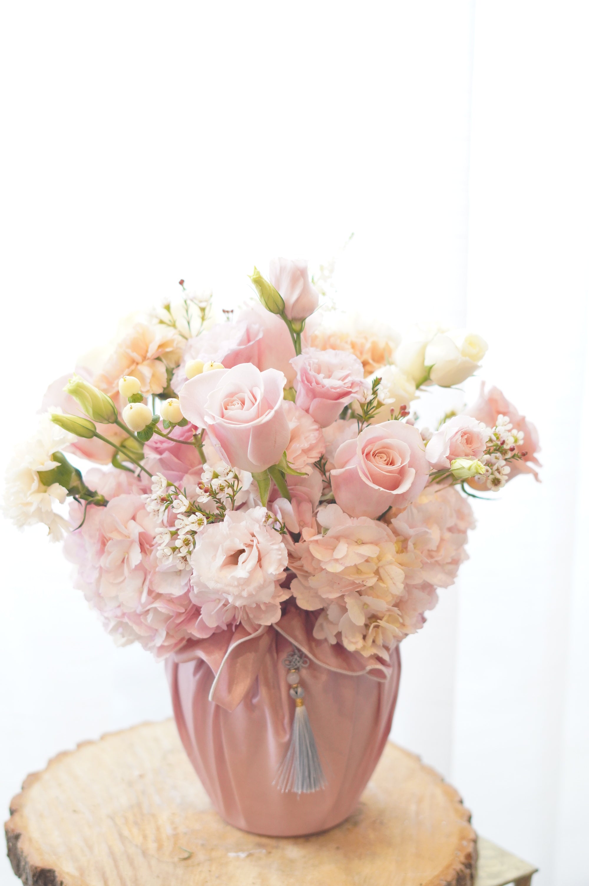 Soft pink and peach Mother's Day floral arrangement with roses and lisianthus in a pink fabric-wrapped vase on a rustic wood slice. Elegant flower delivery in Toronto and Oakville by Moon's Flowers.