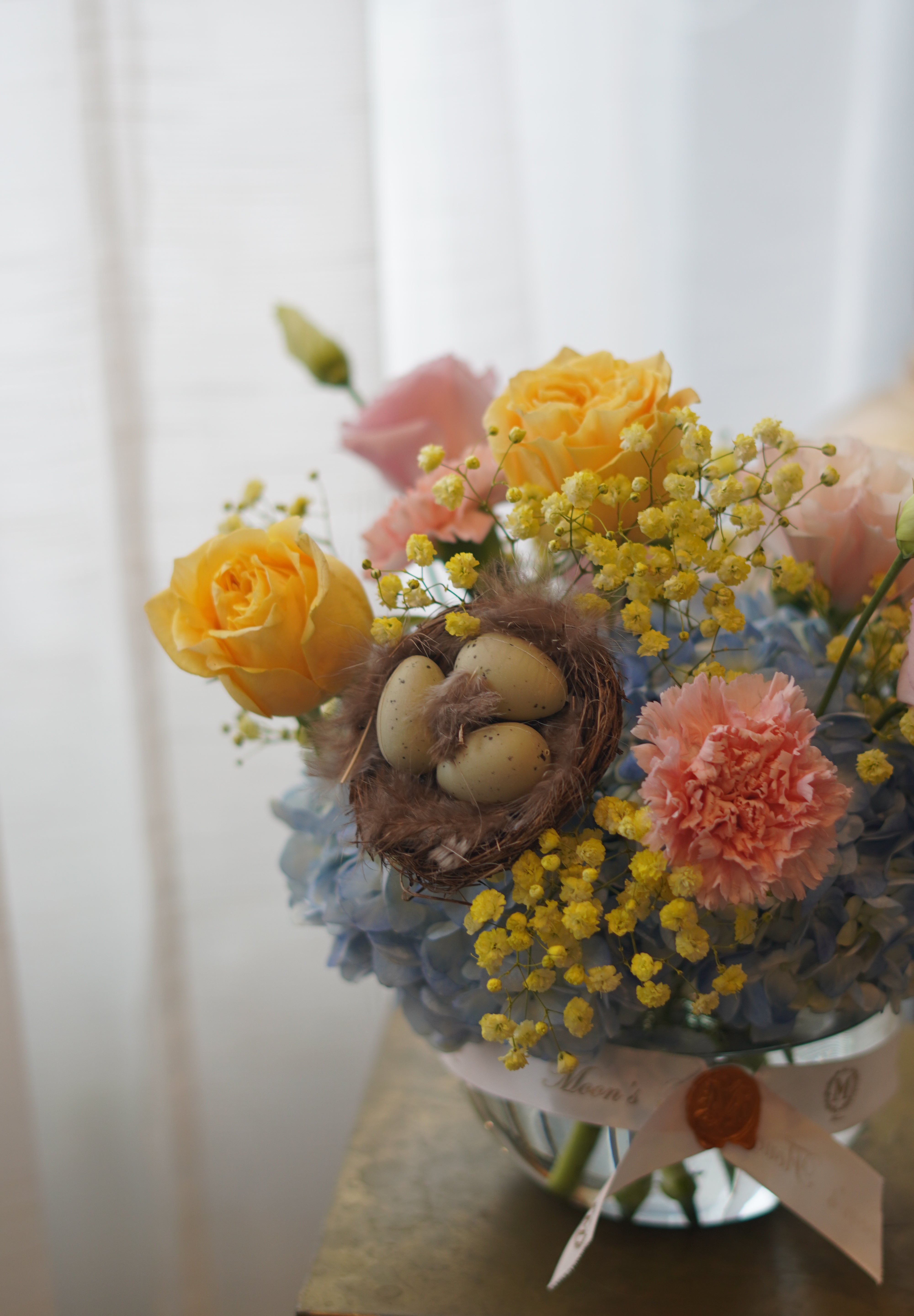 Spring pastel flower arrangement with hydrangeas, roses, mimosa, and decorative nest in glass bowl