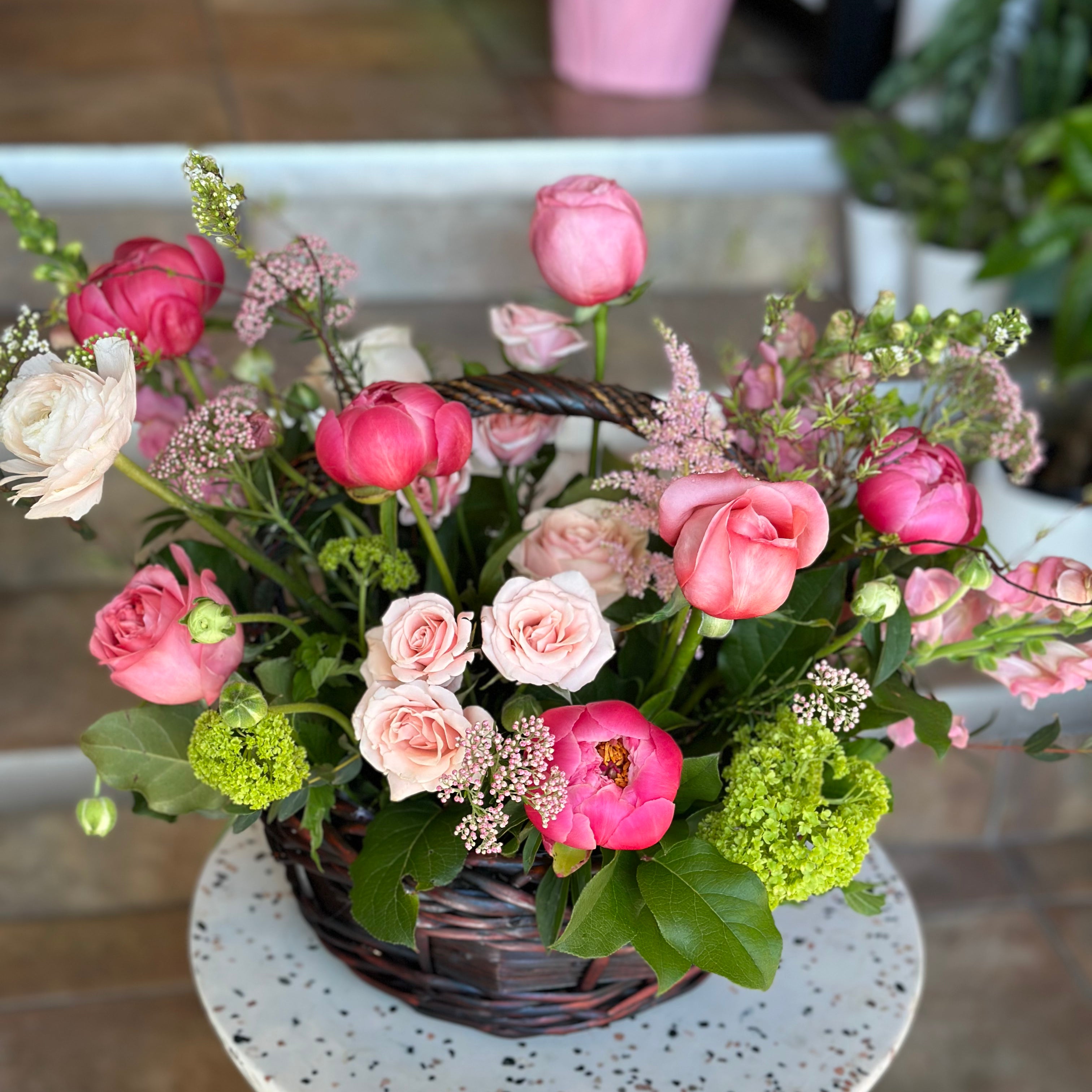 Top-down view of the 'Flowers in a Basket' featuring a lush mix of pink roses, bright peonies, and green hydrangeas on a white speckled stool. A charming Mother's Day floral arrangement by Moon's Flowers Toronto