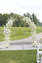 A white floral arch decorated with dense clusters of white flowers, set outdoors on a paved path over a green lawn with trees in the background.