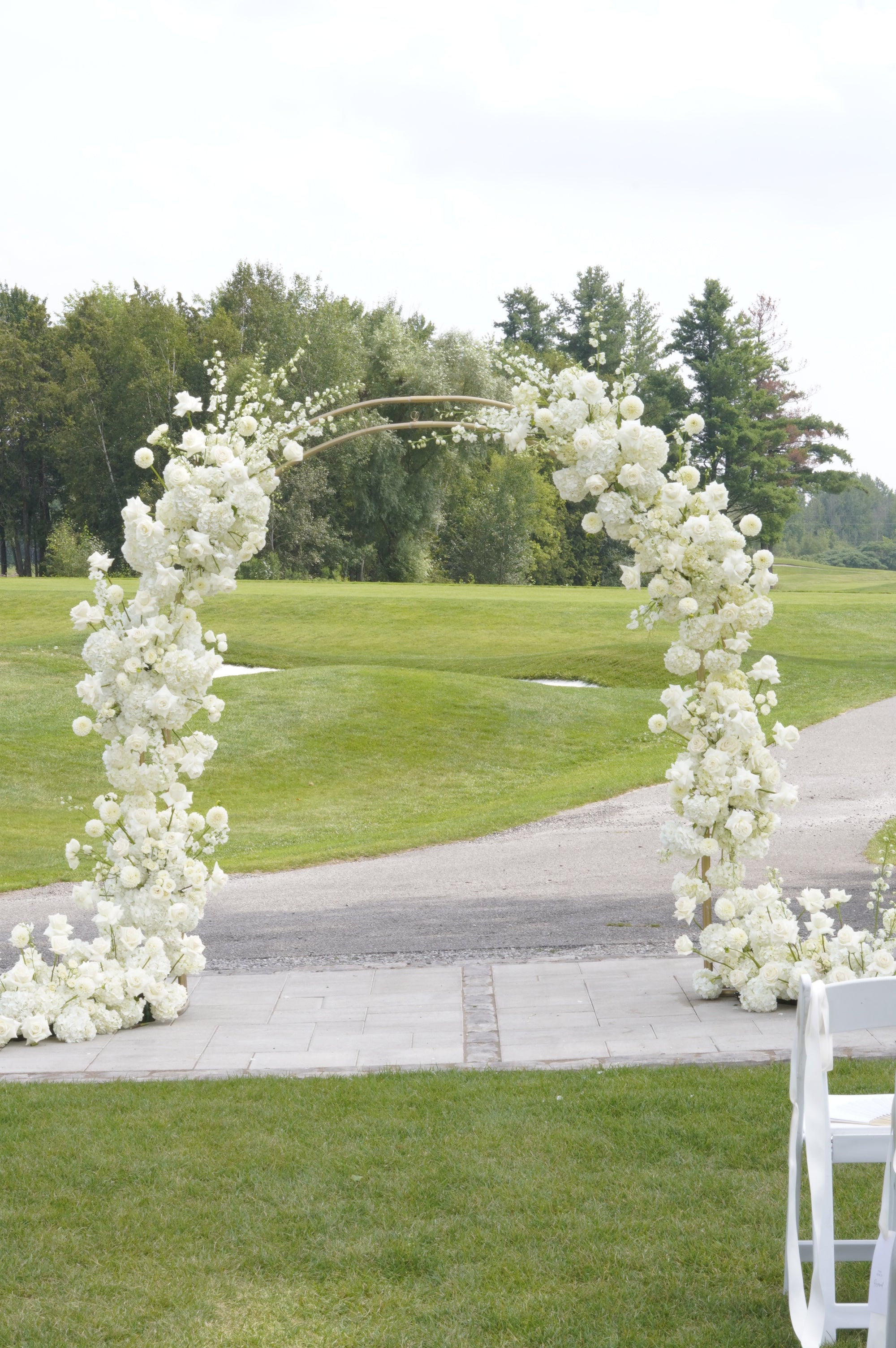 A white floral arch decorated with dense clusters of white flowers, set outdoors on a paved path over a green lawn with trees in the background.