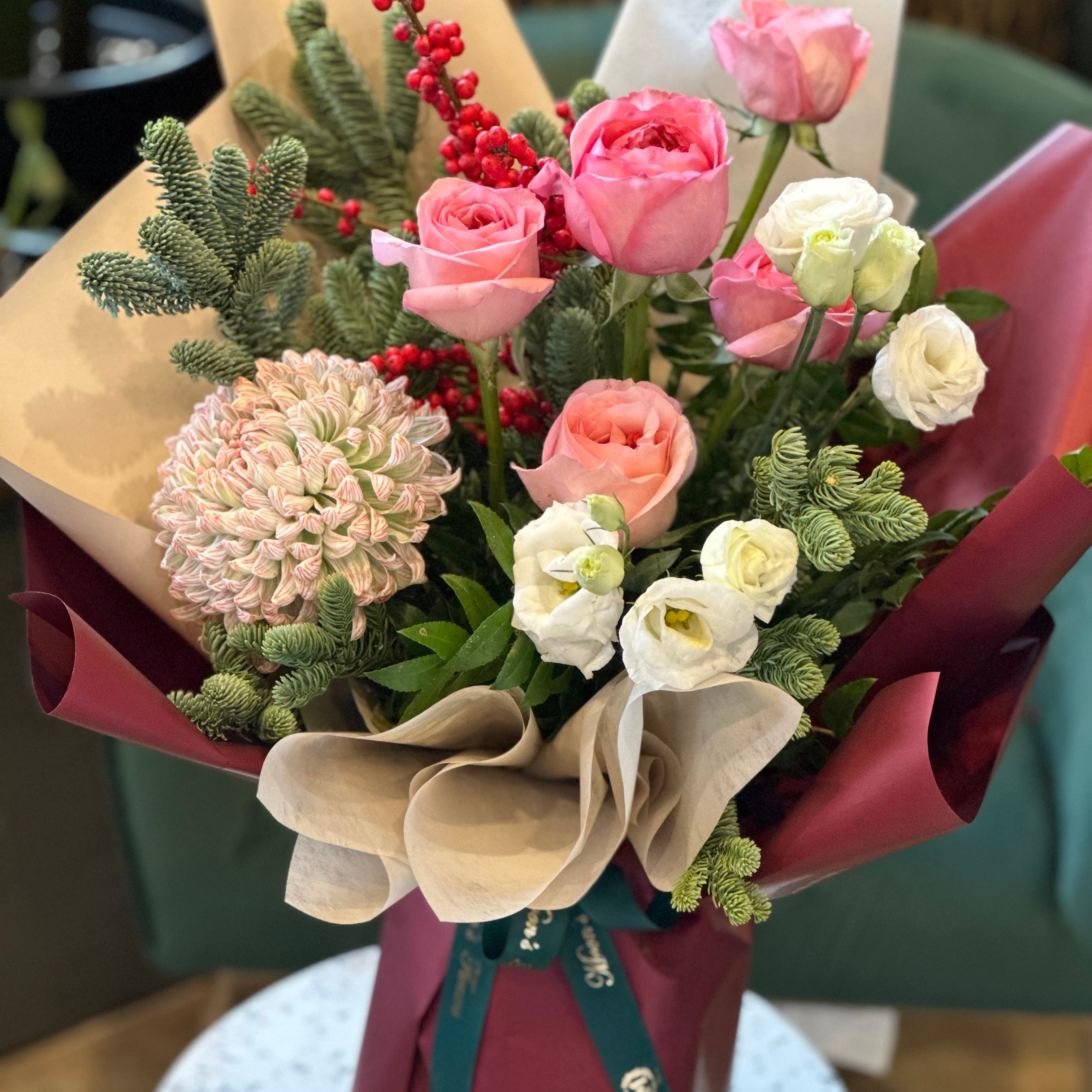 A hand-tied bouquet featuring a mix of pink, white, and red flowers, with green winter foliage and decorative berries, wrapped with brown and white paper.