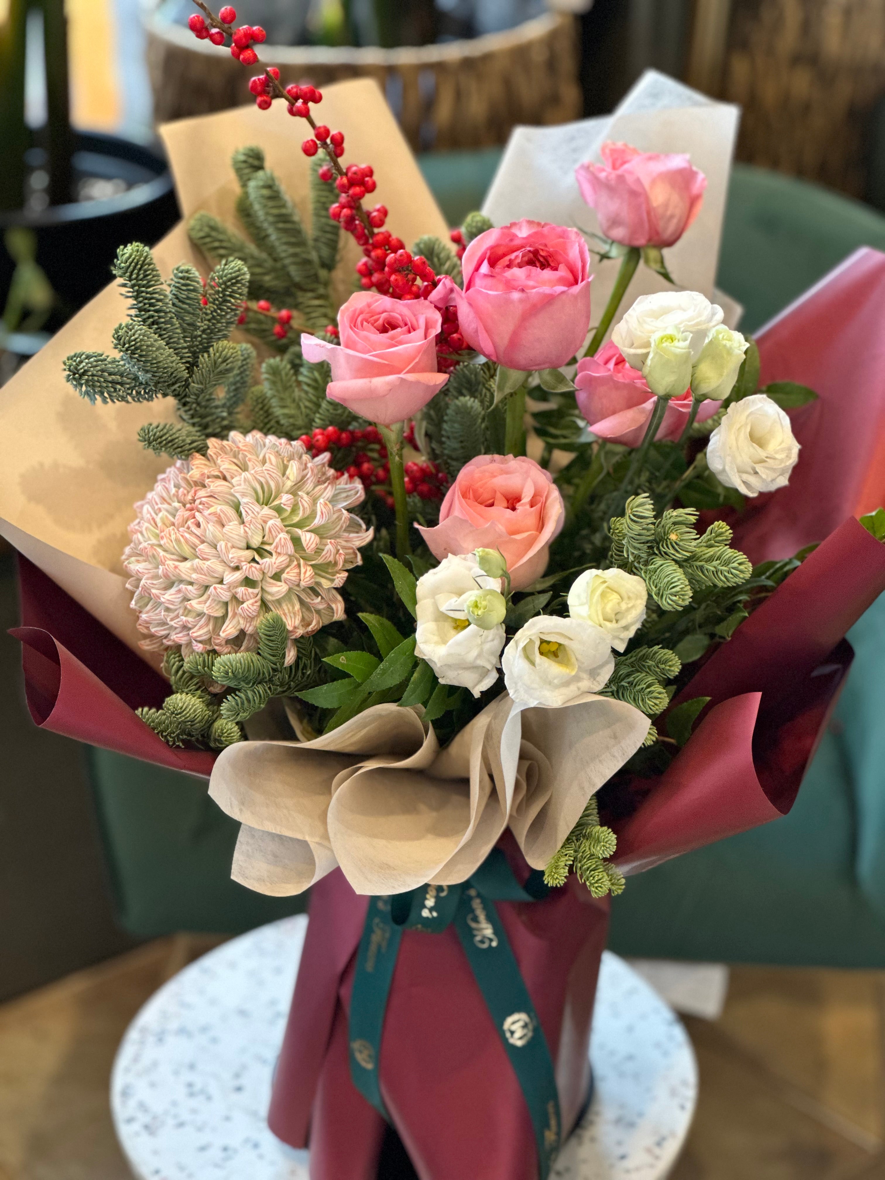 A hand-tied bouquet featuring a mix of pink, white, and red flowers, with green winter foliage and decorative berries, wrapped with brown and white paper.