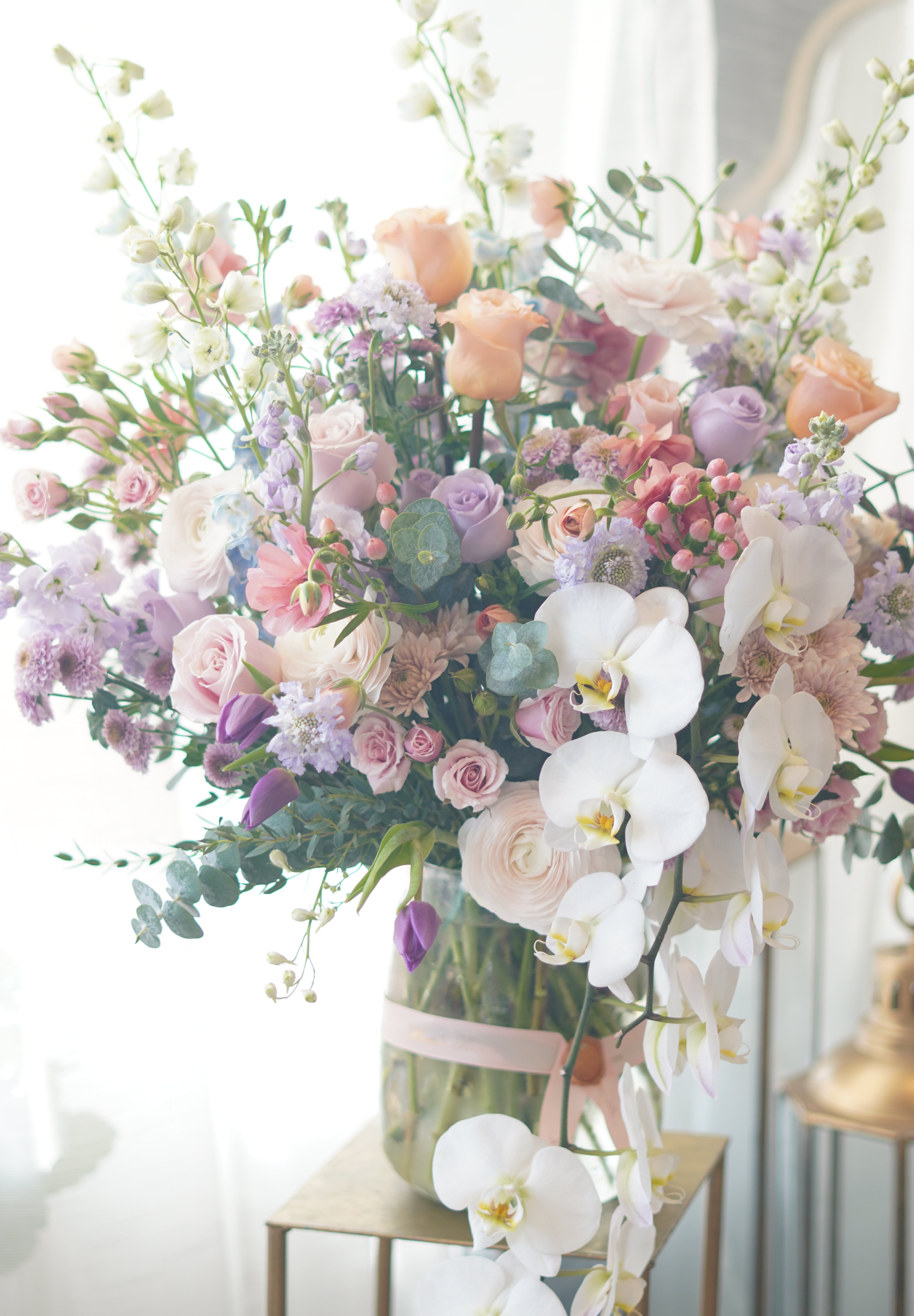 A luxury floral arrangement featuring purple orchids, pink roses, and white ranunculus in a clear glass vase.