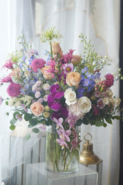 A luxury floral arrangement featuring purple orchids, pink roses, and white ranunculus in a clear glass vase.