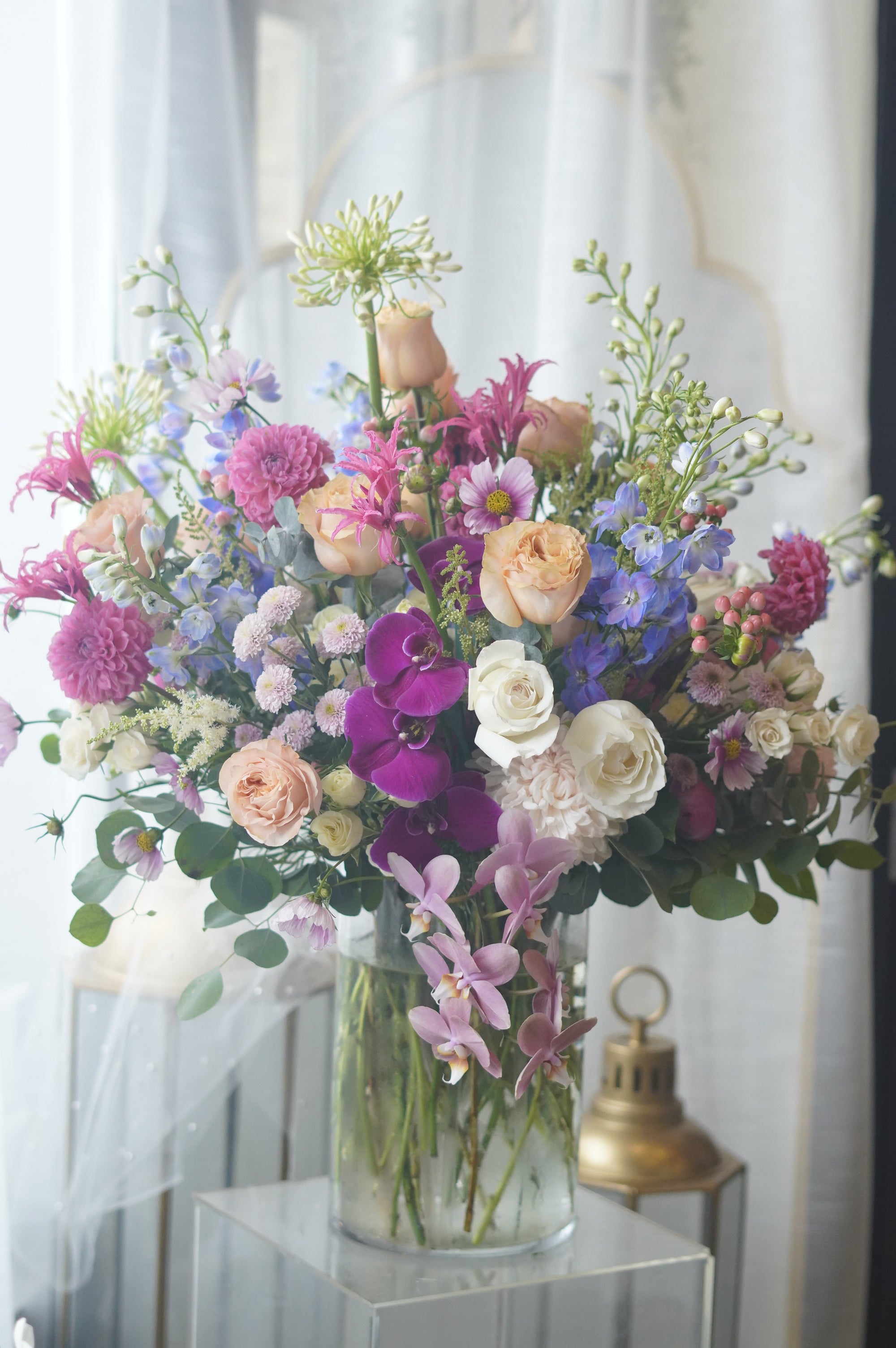 A luxury floral arrangement featuring purple orchids, pink roses, and white ranunculus in a clear glass vase.