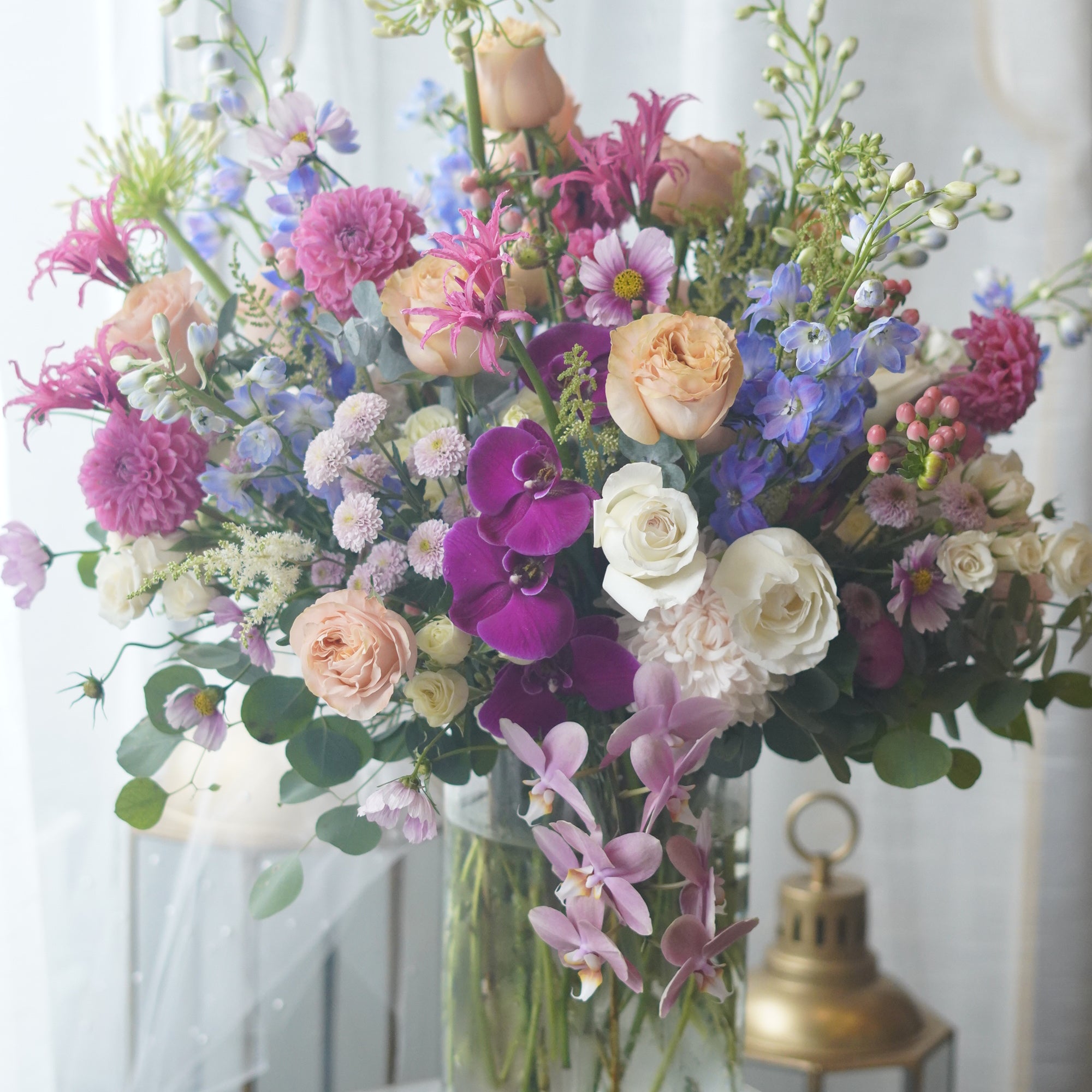 Deluxe luxury flower arrangement with vibrant purple orchids, peach and pink roses, blue delphinium, and fresh greenery in a glass vase.