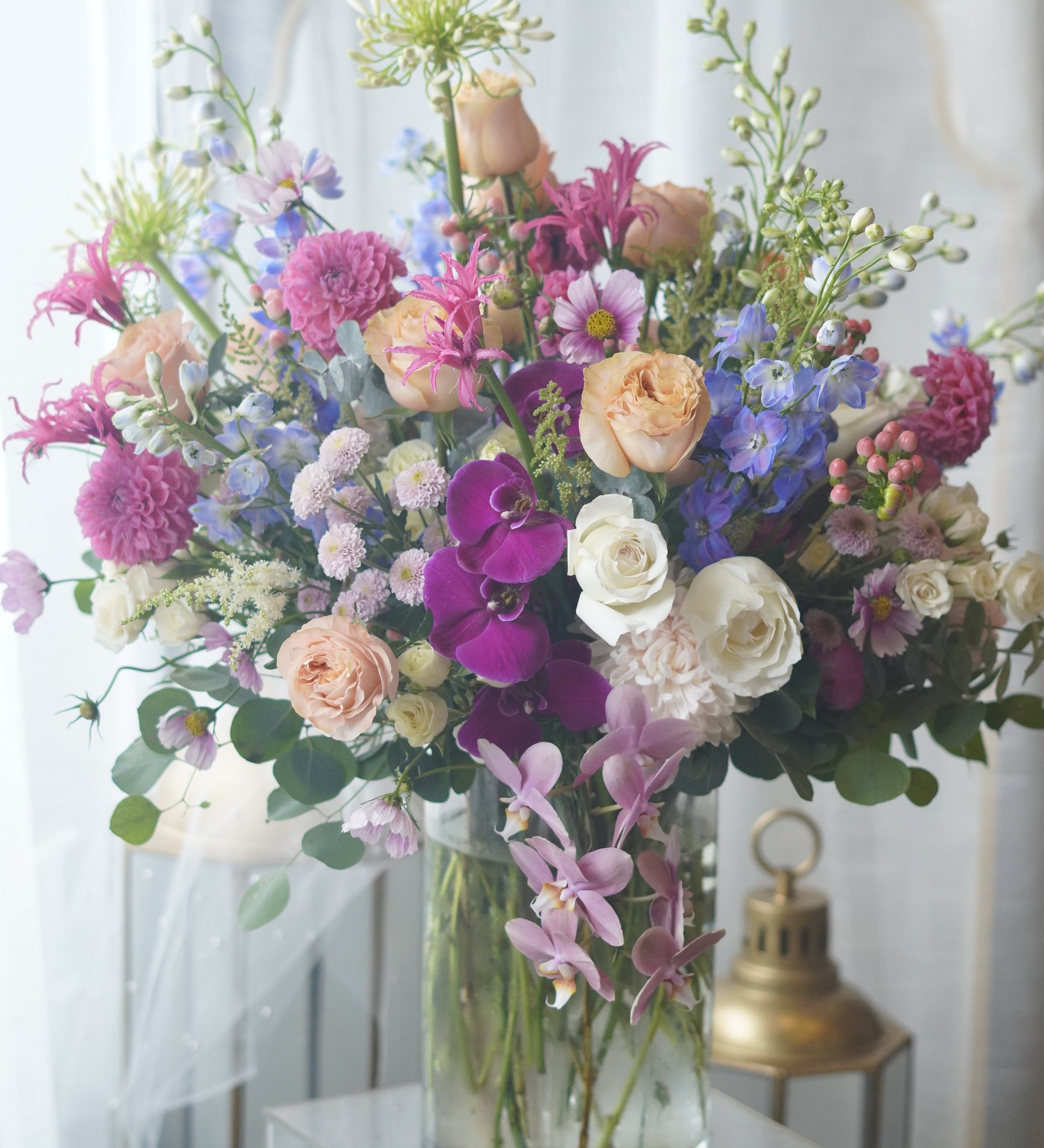 Deluxe luxury flower arrangement with vibrant purple orchids, peach and pink roses, blue delphinium, and fresh greenery in a glass vase.