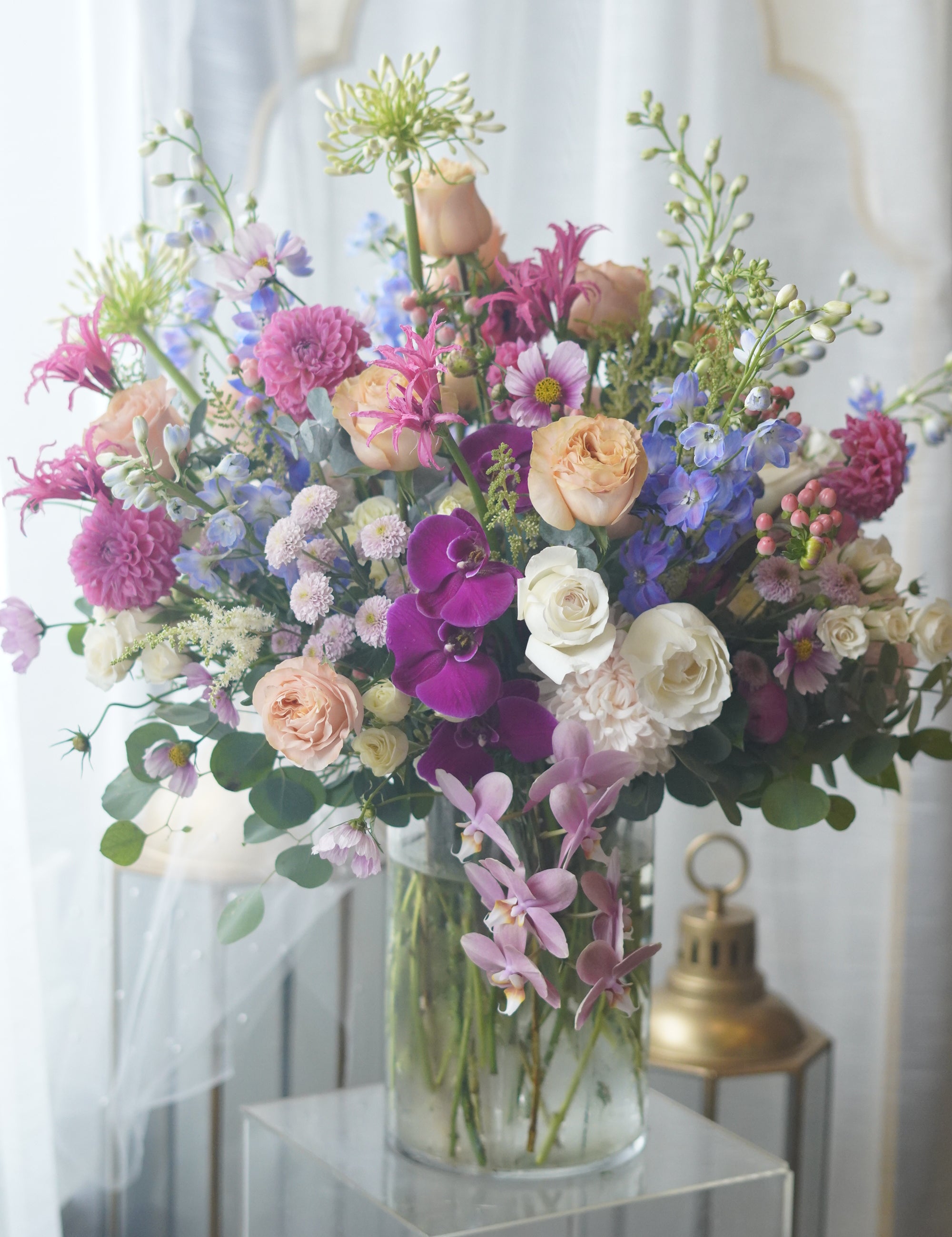Deluxe luxury flower arrangement with vibrant purple orchids, peach and pink roses, blue delphinium, and fresh greenery in a glass vase.