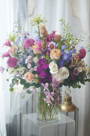Deluxe luxury flower arrangement with vibrant purple orchids, peach and pink roses, blue delphinium, and fresh greenery in a glass vase.