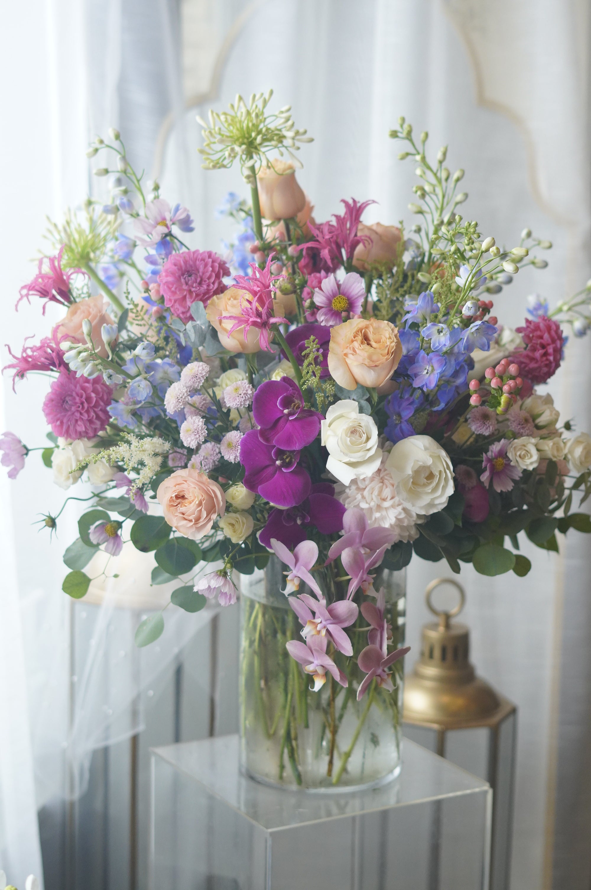 Deluxe luxury flower arrangement with vibrant purple orchids, peach and pink roses, blue delphinium, and fresh greenery in a glass vase.