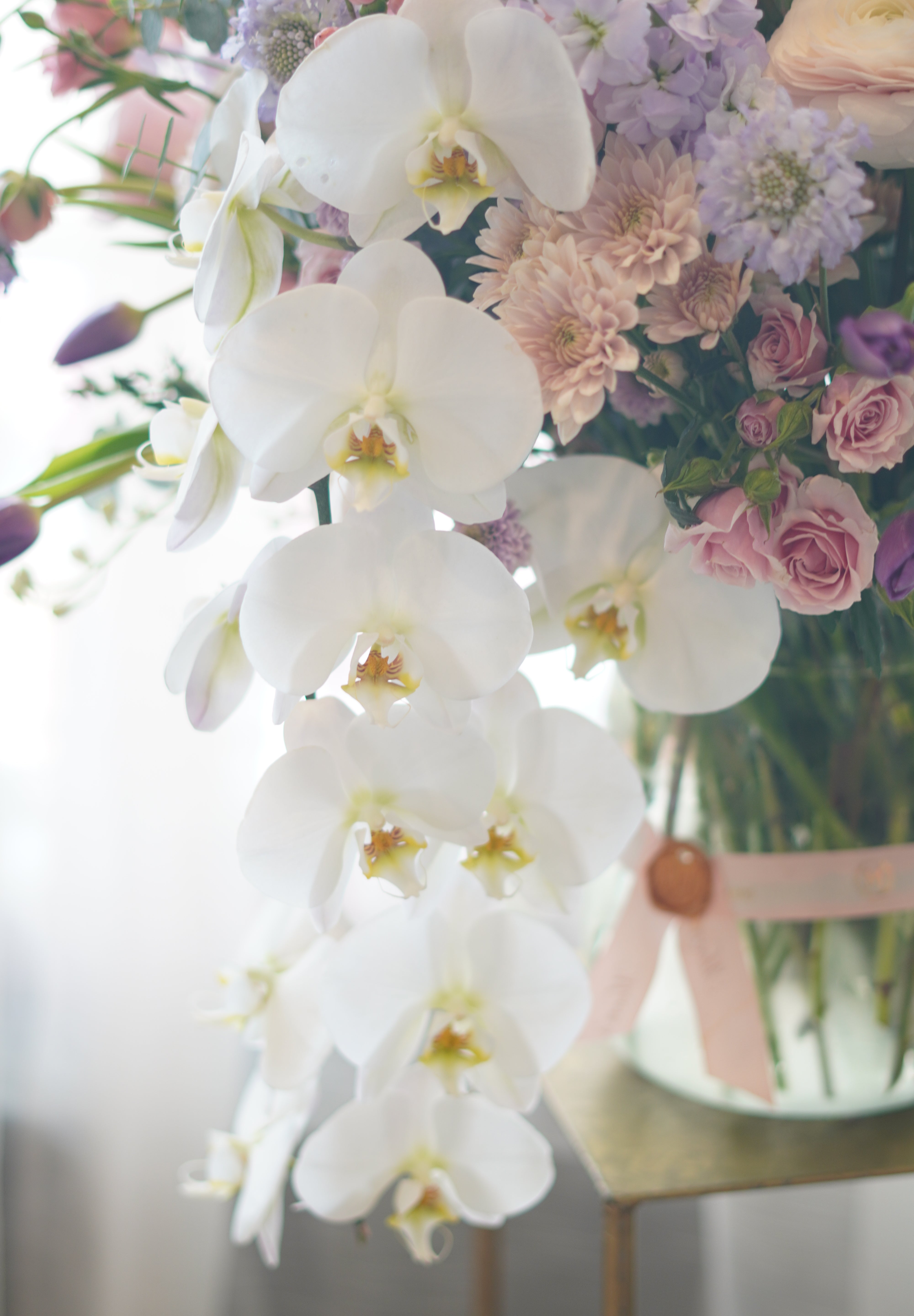 A large, elegant flower bouquet in a clear glass vase resting on an acrylic stand, filled with purple orchids, pink dahlias, peach roses, and mixed greenery against a sheer white backdrop.