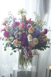 A large, elegant flower bouquet in a clear glass vase resting on an acrylic stand, filled with purple orchids, pink dahlias, peach roses, and mixed greenery against a sheer white backdrop.