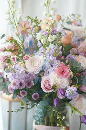 A large, elegant flower bouquet in a clear glass vase resting on an acrylic stand, filled with purple orchids, pink dahlias, peach roses, and mixed greenery against a sheer white backdrop.