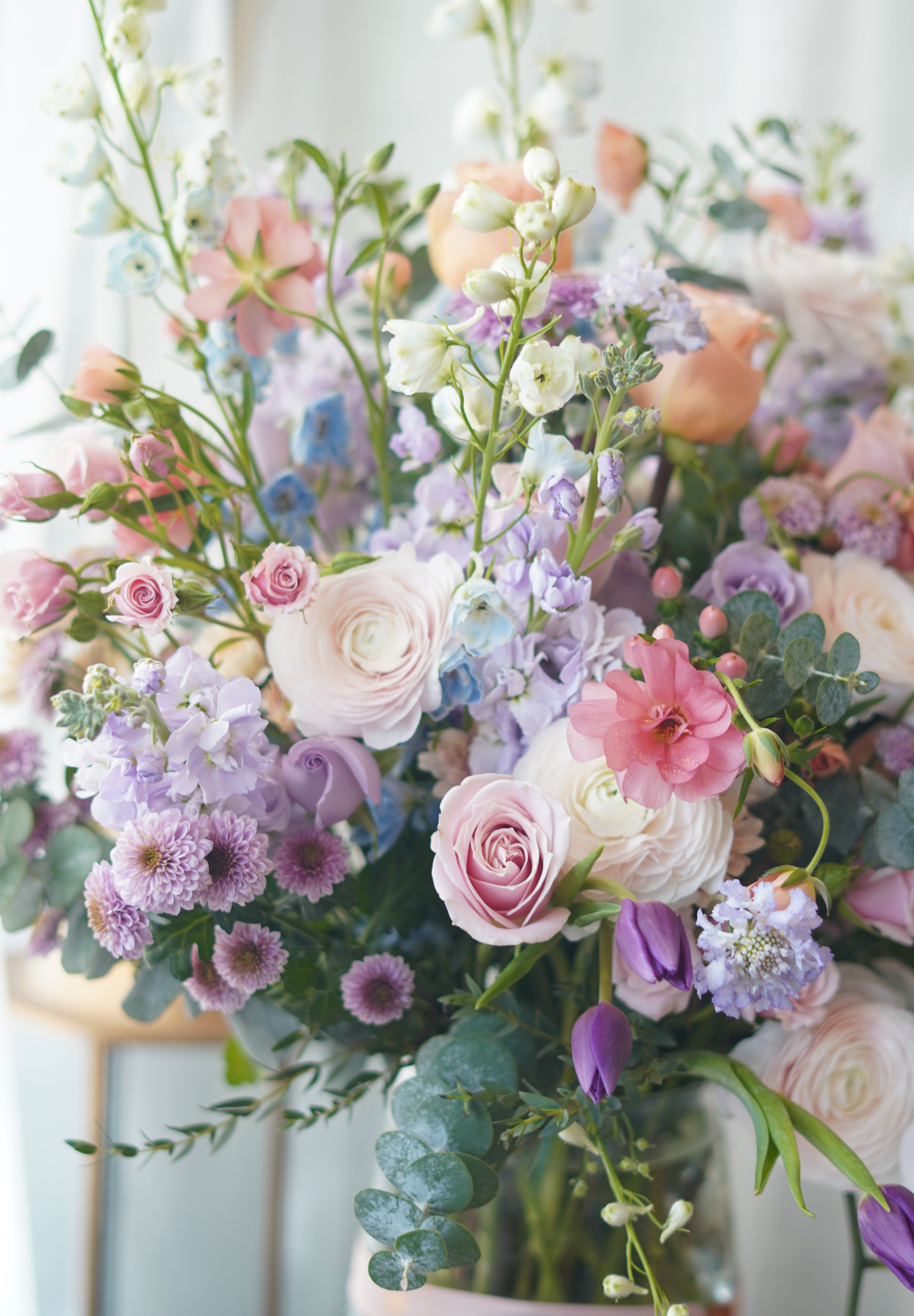 A large, elegant flower bouquet in a clear glass vase resting on an acrylic stand, filled with purple orchids, pink dahlias, peach roses, and mixed greenery against a sheer white backdrop.