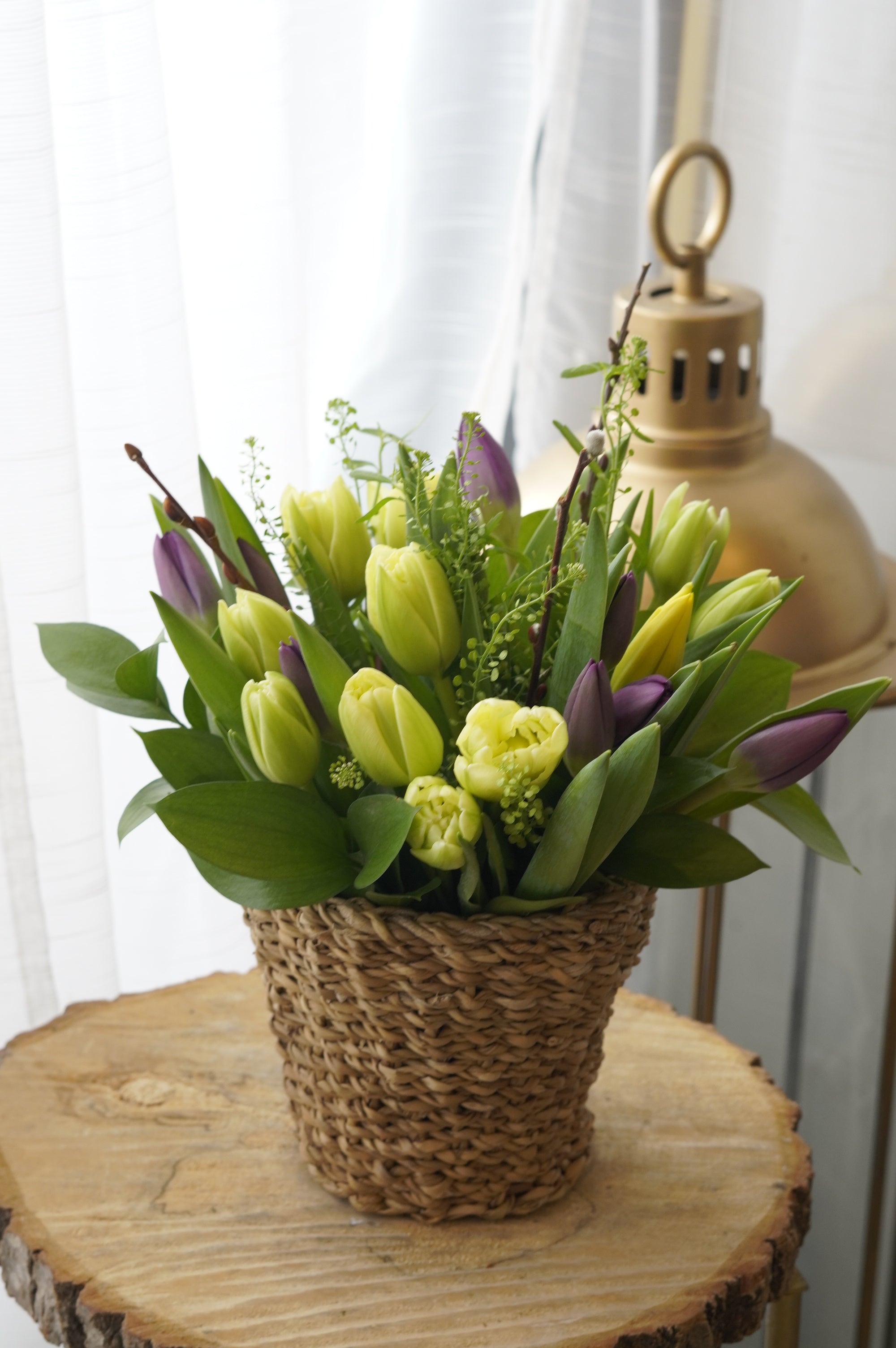 A warm and cheerful seasonal flower basket featuring various shades of tulip and delicate spring catkins, presented in a woven planter for a rustic touch
