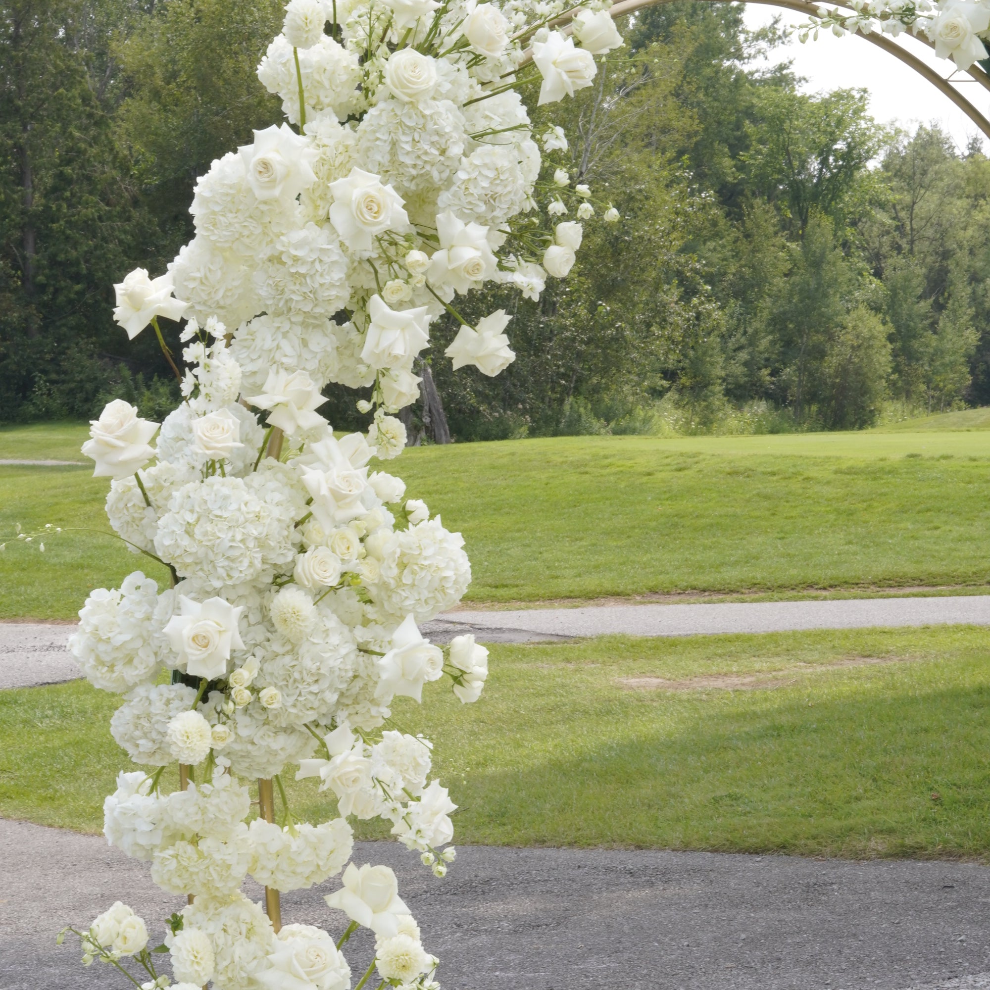 A tall floral arch decoration covered in white flowers, set outdoors on a paved path with green grass and trees in the background.