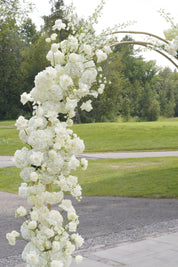 A tall floral arch decoration covered in white flowers, set outdoors on a paved path with green grass and trees in the background.