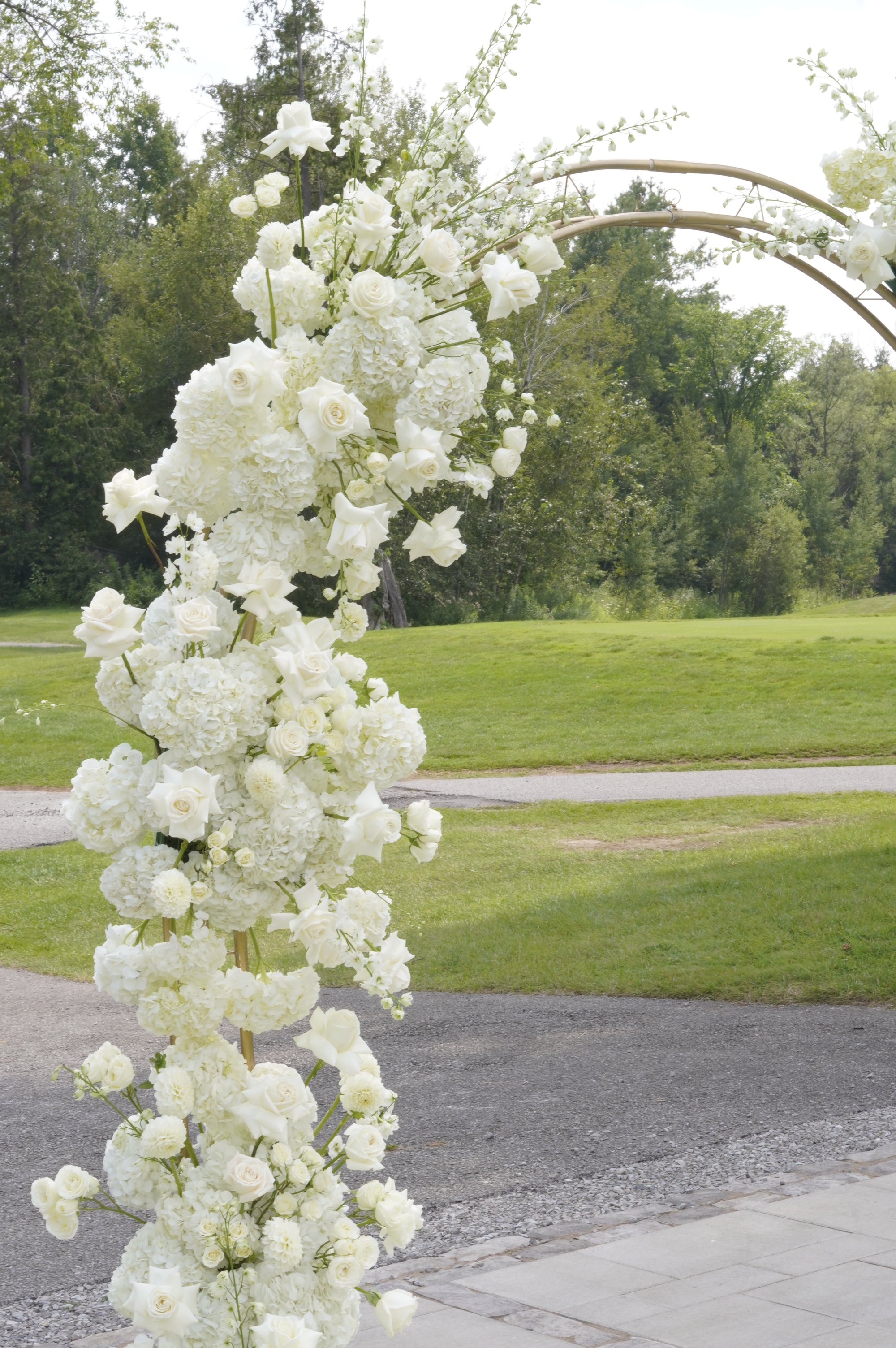 A tall floral arch decoration covered in white flowers, set outdoors on a paved path with green grass and trees in the background.