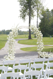 White folding chairs arranged in rows facing a floral archway decorated with white flowers, set outdoors on a grassy golf course with trees in the background.