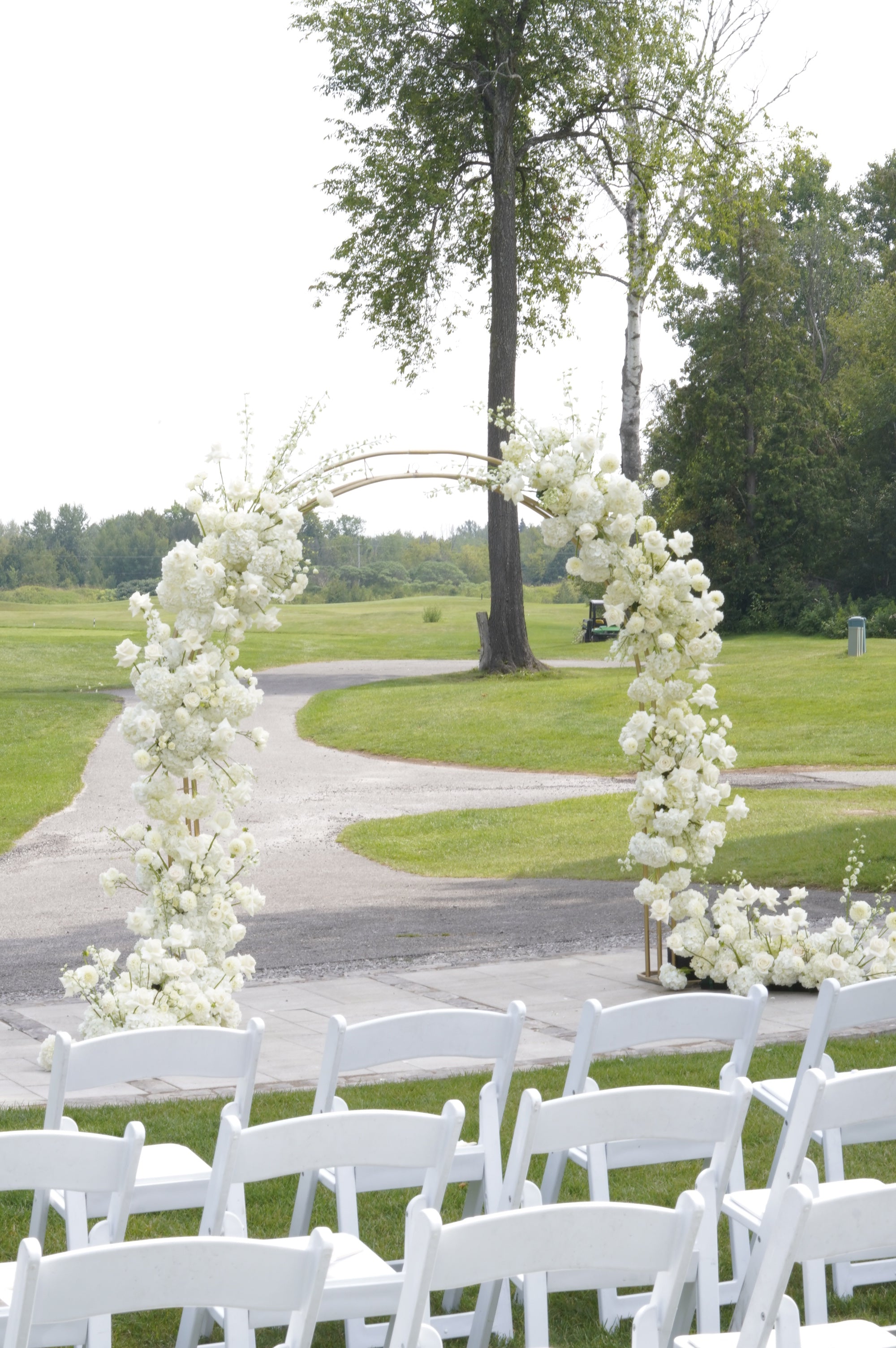 White folding chairs arranged in rows facing a floral archway decorated with white flowers, set outdoors on a grassy golf course with trees in the background.