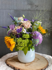 • A cheerful Mother's Day floral arrangement featuring vibrant sunflowers, purple seasonal blooms, and peach roses in a white ceramic vase.
• Displayed on a rustic wooden slice, this Bright and Beautiful bouquet by Moon's Flowers Toronto is perfect for home decor and gifting