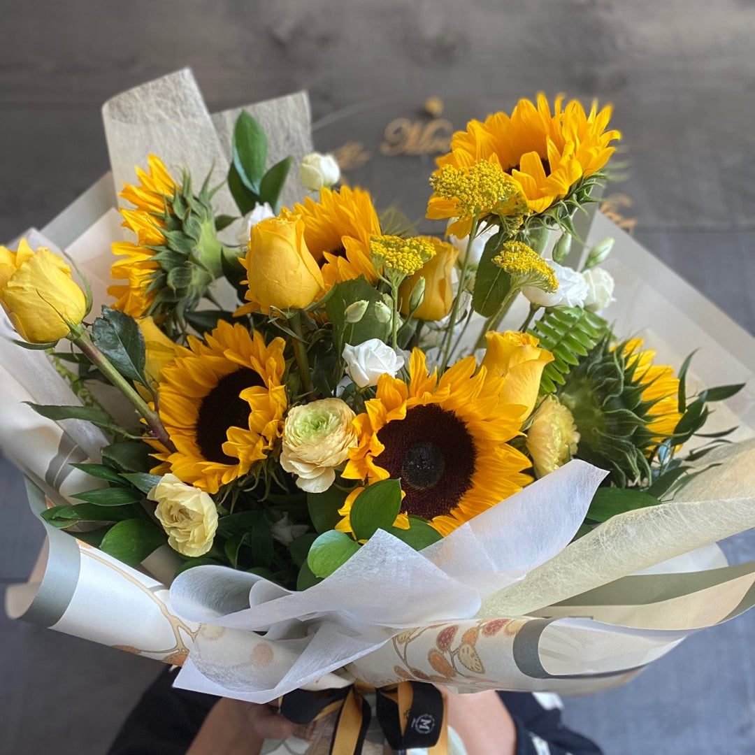 A vibrant bouquet of bright yellow sunflowers, yellow roses, and white accent flowers, elegantly wrapped in white and patterned paper with a black and gold ribbon.