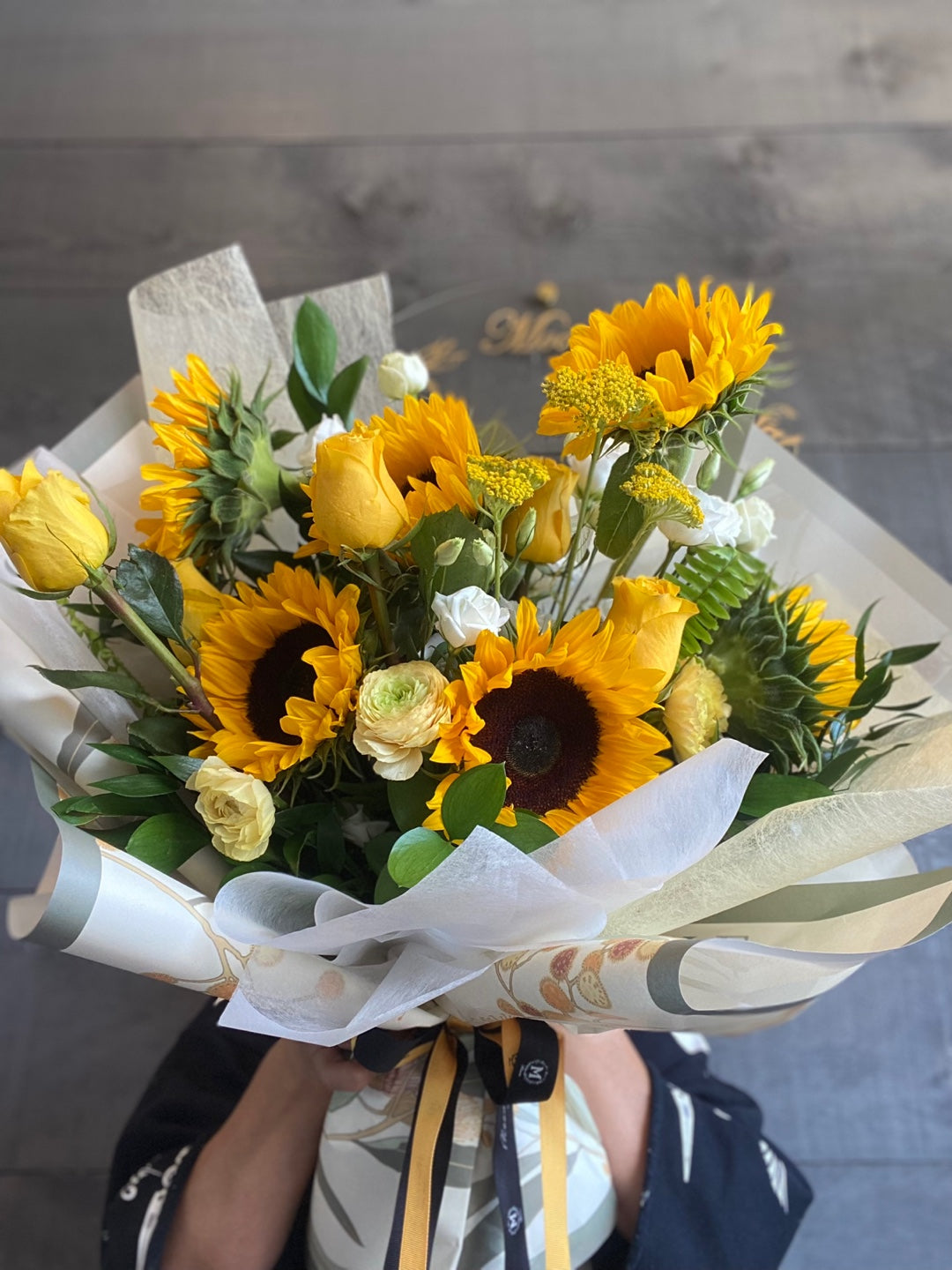 A vibrant bouquet of bright yellow sunflowers, yellow roses, and white accent flowers, elegantly wrapped in white and patterned paper with a black and gold ribbon.