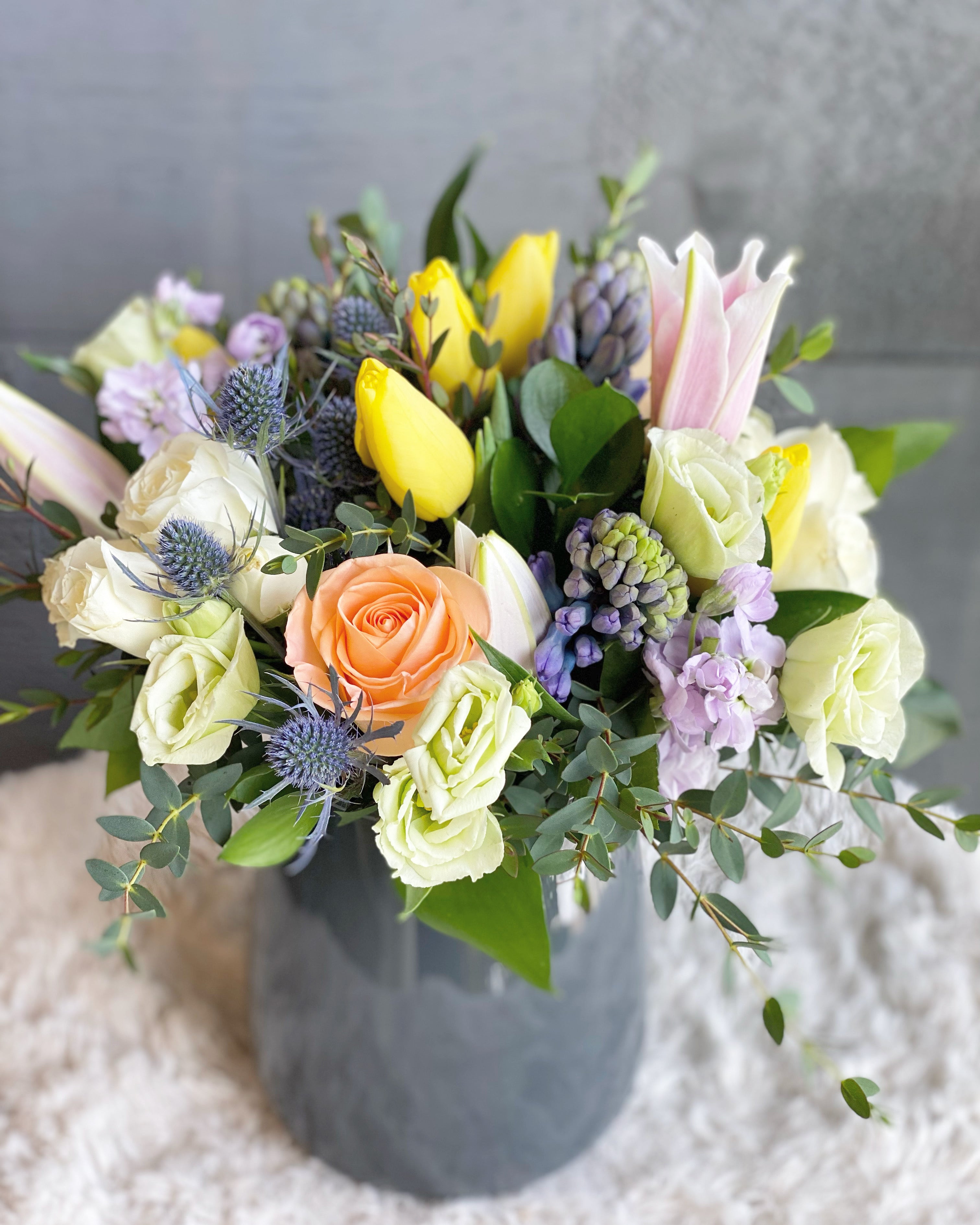Vibrant mixed floral arrangement in a grey vase with peach roses, yellow tulips, purple hyacinths, and thistles. Perfect for local flower delivery